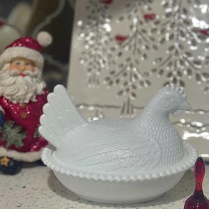May include: A white ceramic hen-shaped dish with a textured surface, designed to hold eggs. The dish has a detailed design and sits on a white base with a beaded edge. A Santa Claus figurine and a decorative plate are in the background.