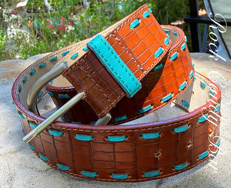 May include: A brown leather belt with a crocodile texture and turquoise stitching. The belt has a silver buckle and a turquoise leather loop. The belt is approximately 1.5 inches wide.