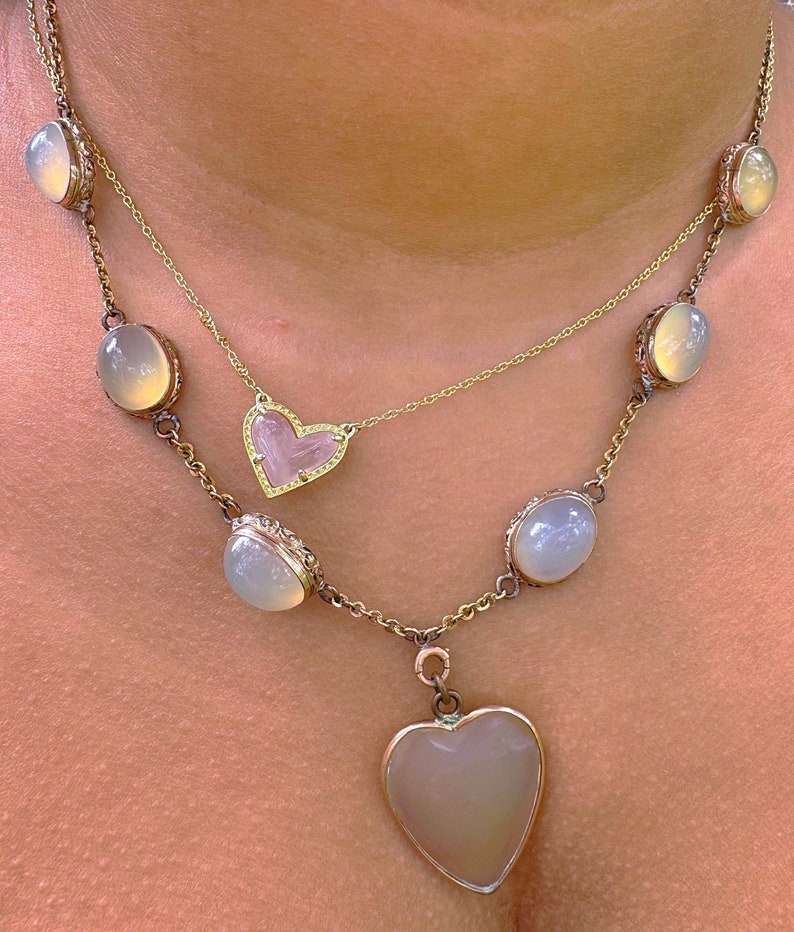 May include: A gold chain necklace with a heart-shaped pendant and oval-shaped stones. The pendant is made of clear quartz with a gold border. The stones are a pale, milky white.