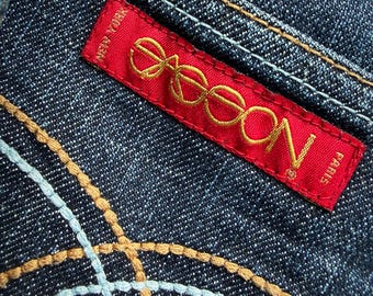 May include: Close-up of dark blue denim fabric with decorative stitching in light blue and tan. A red label with gold lettering reads "eeoon" and "New York Paris".