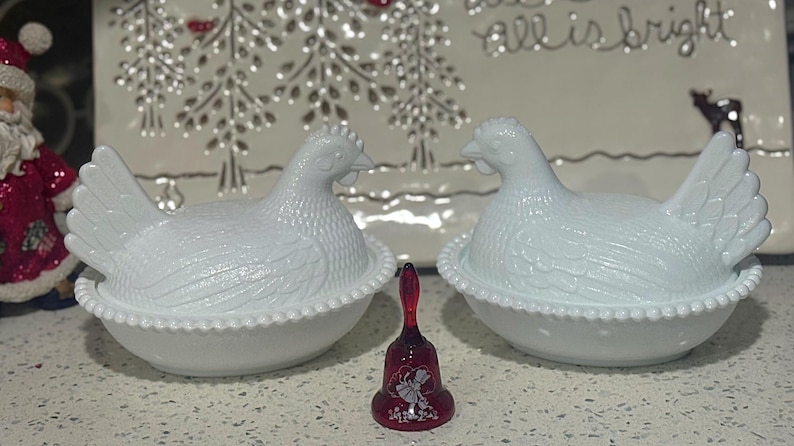 May include: Two white ceramic hen-shaped covered dishes with textured details, sitting on a white surface. A small, red bell with a floral design is between them. A decorative plate with the words "all is bright" is in the background.