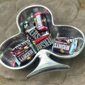 May include: A silver clover-shaped dish filled with assorted Hershey's chocolate bars. The bars are wrapped in foil and feature the Hershey's logo and the words "milk chocolate" and "special".