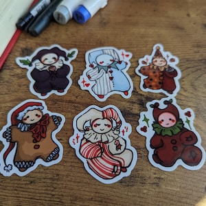 May include: A collection of six whimsical sticker designs featuring cartoon-style jesters in various outfits. The stickers showcase a range of colors, including red, brown, blue, and white, with decorative elements like hearts and holly. The stickers are on a wooden surface.