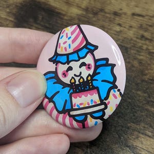 May include: A round, pink button featuring a cartoon clown holding a birthday cake with three lit candles. The clown wears a party hat and a blue ruffled collar. The button is decorated with colorful confetti.