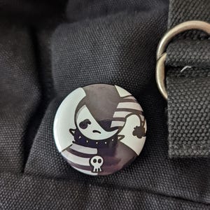 May include: A round button pin with a black and white cartoon design. The design features a crescent moon with a face, a striped hat, and a skull. The pin is set against a black fabric background.
