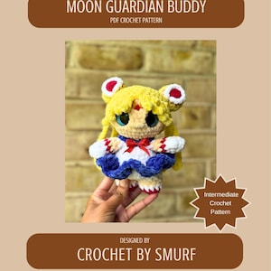 Puede incluir: Muñeca de ganchillo con cabello amarillo, ojos azules y un lazo rojo. La muñeca lleva un vestido azul y blanco. El texto de la imagen dice "Moon Guardian Buddy" y "Crochet by Smurf". El patrón es un patrón de ganchillo intermedio.
