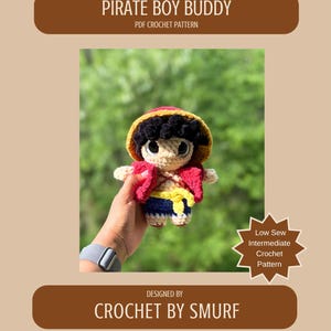 May include: A crocheted doll in the image of a pirate, with a red jacket, yellow sash, and blue shorts. The doll has a straw hat and black hair. The text on the image says "PIRATE BOY BUDDY" and "CROCHET BY SMURF".