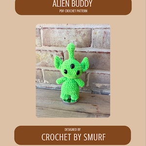 May include: A green crocheted alien with three black eyes, small arms, and a tall head. The text "ALIEN BUDDY PDF CROCHET PATTERN" is at the top, and "DESIGNED BY CROCHET BY SMURF" is at the bottom. The alien is on a wooden surface.