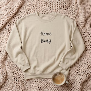 May include: A beige crewneck sweatshirt featuring the text Home Body in a black cursive font centered on the chest. The garment has long sleeves with ribbed cuffs and a ribbed neckline, presented in a simple, clean design for casual wear.