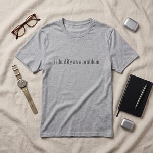 May include: A heather grey short-sleeved t-shirt featuring the black text "i identify as a problem" printed across the chest. The shirt is displayed alongside brown-rimmed glasses, a wristwatch with a tan strap, a black notebook, a pen, and earbuds.