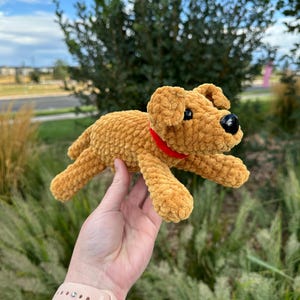 May include: A crocheted plush toy dog in a brown color with a red collar. The dog is lying on its belly with its legs outstretched.