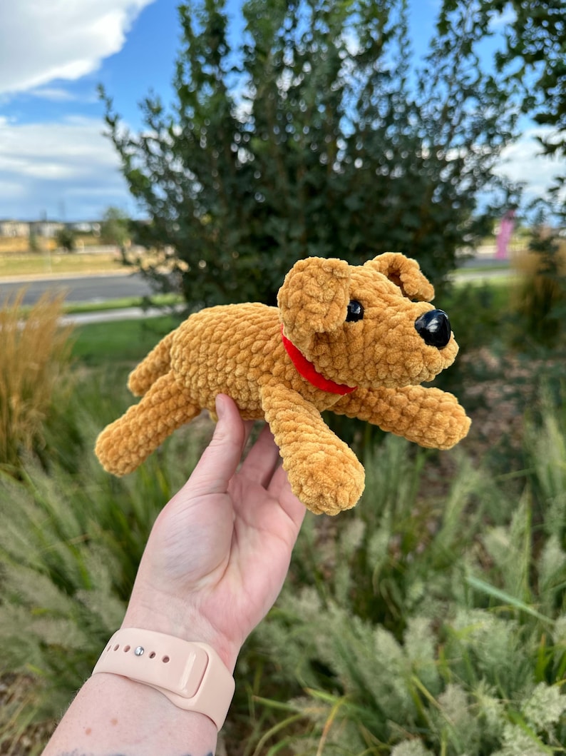 May include: A crocheted plush toy dog in a brown color with a red collar. The dog is lying on its belly with its legs outstretched.