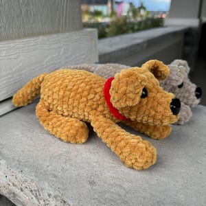 May include: Two crocheted plush dog toys. One is brown with a red collar and the other is gray. Both are lying on a gray stone surface.