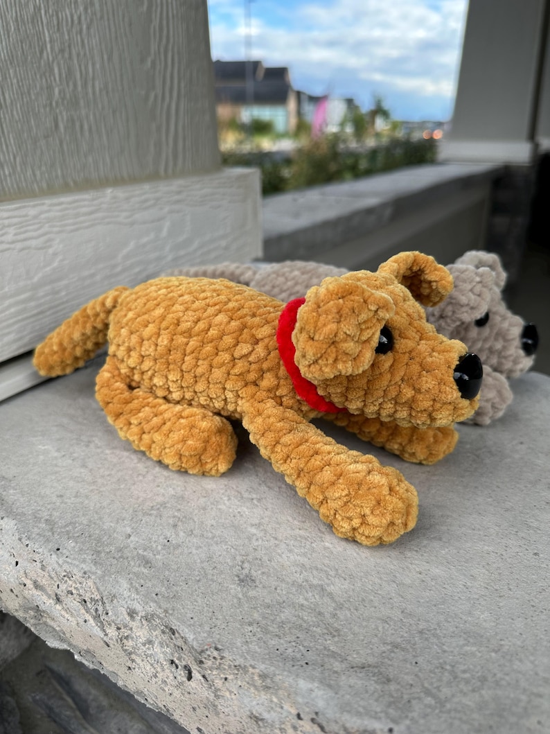 May include: Two crocheted plush dog toys. One is brown with a red collar and the other is gray. Both are lying on a gray stone surface.