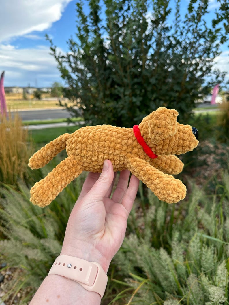 May include: A crocheted plush dog toy in a light brown color with a red collar. The dog is lying on its side with its head turned to the right.