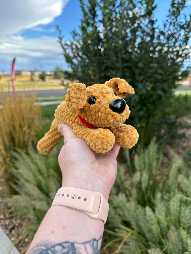 May include: A crocheted plush dog toy with brown fur, black eyes, and a red collar. The toy is being held in a person's hand.