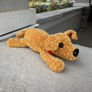 May include: A crocheted brown dog plush toy with black eyes and a red bow. The toy is lying on a gray concrete surface.
