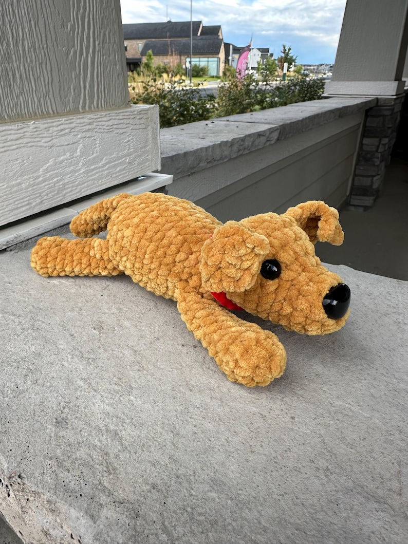 May include: A crocheted brown dog plush toy with black eyes and a red bow. The toy is lying on a gray concrete surface.