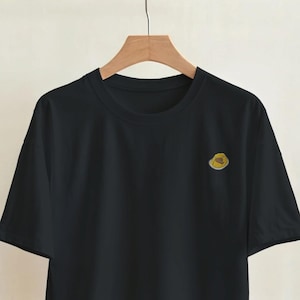 May include: A black short-sleeved t-shirt hanging on a wooden hanger. The shirt features a small, circular yellow embroidered graphic on the upper left chest area. The garment has a standard crew neckline and a straight hemline design.