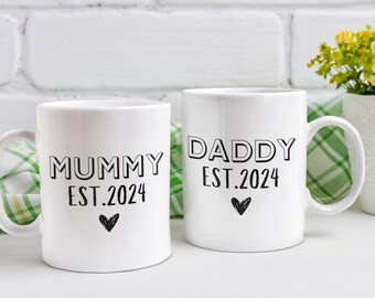Puede incluir: Dos tazas de cerámica blanca con texto negro. La taza de la izquierda dice "Mummy Est. 2024" con un pequeño corazón. La taza de la derecha dice "Daddy Est. 2024" con un pequeño corazón.