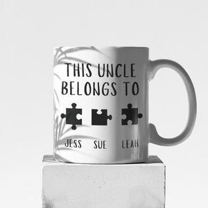 Puede incluir: Taza de cerámica blanca con texto negro que dice "This Uncle Belongs To" y tres piezas de rompecabezas con los nombres "Jess", "Sue" y "Leah" debajo.