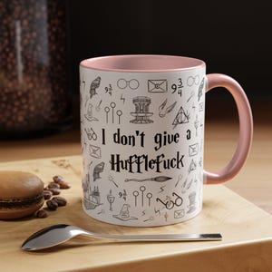 May include: White ceramic mug with a pink interior and handle. The mug features black line art and the text "I don't give a Hufflefuck". A spoon and a macaron are in the foreground, resting on a wooden board.