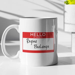Puede incluir: Taza de cerámica blanca con una etiqueta de nombre roja que dice "Hola, mi nombre es Regina Phalange".
