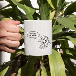 以下が含まれることがあります： 白いセラミック製のマグカップ。黒と白の鶏のイラストが描かれており、吹き出しには「私は鳥語に堪能です」と書かれています。