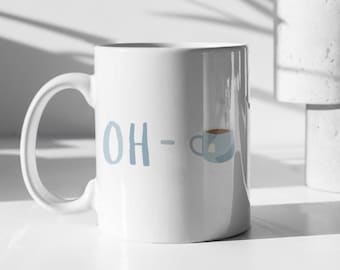 May include: White ceramic mug with the text "OH -" in light blue. A small blue teacup graphic with tea and a teabag is next to the text. The mug has a large handle and is on a white surface.