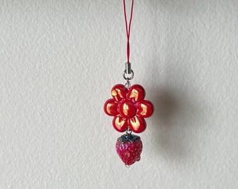 May include: A red flower charm with a red strawberry charm hanging from it. The charms are attached to a red string with a silver clip.