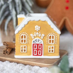 Peut inclure: Un photophore en forme de maison en pain d'épices en céramique, avec un extérieur jaune, des détails de glaçage blanc et une porte rouge. La maison a des fenêtres. Des bâtons de cannelle sont sur un plateau en bois. Décoration de Noël.