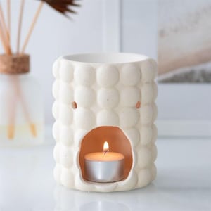 Peut inclure: Diffuseur d'huiles essentielles en céramique crème, avec un motif texturé en forme de bulles. Il présente une ouverture frontale avec une bougie chauffe-plat allumée. Un flacon en verre avec des roseaux est en arrière-plan.