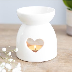 Peut inclure: Un diffuseur d'huiles essentielles en céramique blanche avec une découpe en forme de cœur. Une bougie chauffe-plat allumée est visible à l'intérieur du diffuseur. Le diffuseur a un bol rond sur le dessus pour l'huile et l'eau. Fond bleu clair et surface en bois.