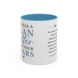 Puede incluir: Una taza de cerámica blanca con interior y borde azules. El exterior presenta texto azul que dice God has a plan better than your fears y un pasaje de Jeremías 29:11, acompañados de pequeños iconos de corazones azules en las superficies.
