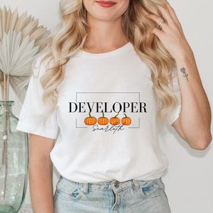 May include: White t-shirt with the text "DEVELOPER" in black lettering and a row of five orange pumpkins with the text "Scarleth" underneath.