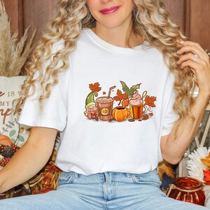 May include: White t-shirt with a fall-themed design featuring a gnome, pumpkin, coffee cups, and autumn leaves. The text "Home is where my pumpkin is" is visible on a cushion in the background.