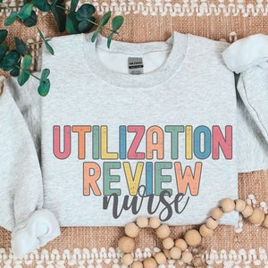 May include: A light gray sweatshirt with the text "UTILIZATION REVIEW nurse" printed in rainbow colors. The text is in a retro, distressed font.