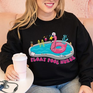 May include: Black sweatshirt with a graphic of a pink flamingo wearing a nurse's hat and sunglasses floating in a pool with a stethoscope. The text "Float Pool Nurse" is printed below the graphic.