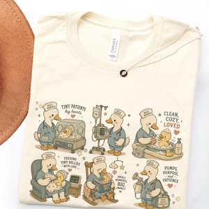 May include: A cream-colored t-shirt featuring a graphic print of ducks dressed as NICU nurses caring for ducklings. The illustrations show medical scenes with text including Tiny Patients Big Hearts, Clean Cozy Loved, and Feeding Tiny Bellies.