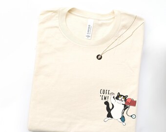 May include: A cream-colored t-shirt featuring a graphic of a black and white cat holding a red blood pressure cuff with the text CUFF 'EM! printed above it. A thin silver-toned necklace with a small circular pendant rests on the shirt neckline.