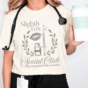 May include: A cream-colored t-shirt featuring a gray graphic with the text "Slightly Tox-ic Social Club The Only Kind Of Toxic You Need." The design includes a lip print, a syringe, a medicine vial, and a laurel wreath frame.