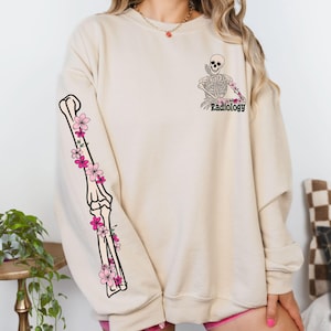 May include: A beige sweatshirt with a skeleton graphic and the word "Radiology" on the front. The left sleeve features a floral design with a skeleton arm.