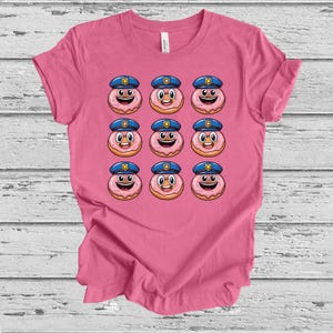 May include: Pink t-shirt with a grid of smiling donut characters wearing police hats. The donuts have pink icing, a smiling face, and a blue police hat. The shirt is made of a soft material.
