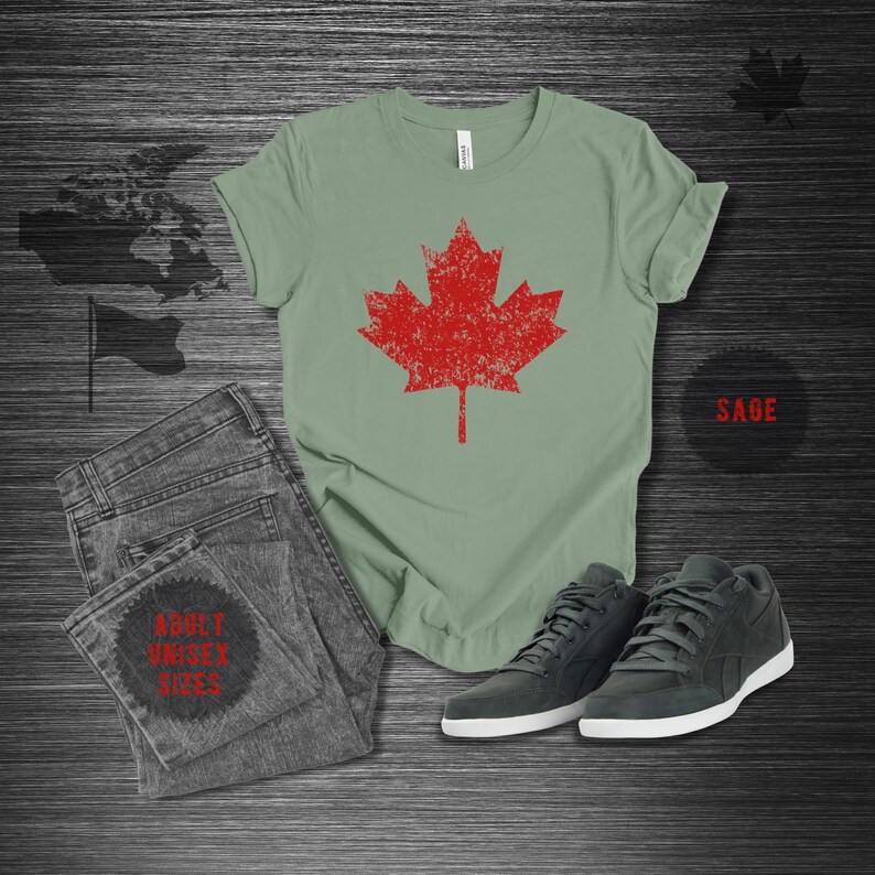 K&ouml;nnte beinhalten: Salbeigr&uuml;nes T-Shirt mit einem roten Ahornblatt-Design im Used-Look. Das Bild zeigt auch graue Jeans, dunkelgraue Sneaker mit wei&szlig;en Sohlen und die Texte "ADULT UNISEX SIZES" und "SAGE".