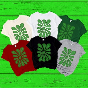 May include: Five t-shirts in different colors with a green Christmas word art design in the shape of a flower. The design includes the words "Merry Christmas" and "Happy Holidays".