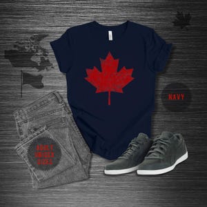 K&ouml;nnte beinhalten: Marineblaues T-Shirt mit einem roten Ahornblatt-Design im Used-Look. Das Bild zeigt eine graue Jeans und dunkelgraue Sneaker mit wei&szlig;en Sohlen. Der Text "NAVY" befindet sich in einem Kreis, und "ADULT UNISEX SIZES" steht auf der Jeans.
