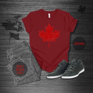 K&ouml;nnte beinhalten: Ein kardinalrotes T-Shirt mit einem roten Ahornblatt-Design im Used-Look. Das Shirt wird mit grauen Jeans und dunkelgrauen Turnschuhen kombiniert. Der Text "ADULT UNISEX SIZES" ist auf der Jeans zu sehen. Das Wort "CARDINAL" ist ebenfalls sichtbar.