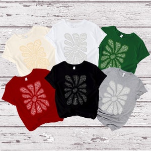 Puede incluir: Seis camisetas de diferentes colores con un diseño de copo de nieve blanco hecho con las palabras "Feliz Navidad". Las camisetas se muestran sobre un fondo de madera blanca.