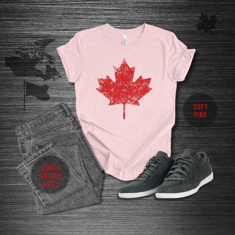 K&ouml;nnte beinhalten: Ein hellrosa T-Shirt mit einem roten Ahornblatt-Design im Used-Look, kombiniert mit grauen Jeans und dunkelgrauen Turnschuhen. Das Shirt ist mit "Soft Pink" und "Adult Unisex Sizes" beschriftet. Eine kanadische Flagge und Karte sind im Hintergrund.