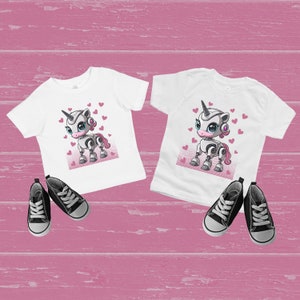 May include: Two white t-shirts with a graphic of a robot unicorn with pink hearts on a white background.
